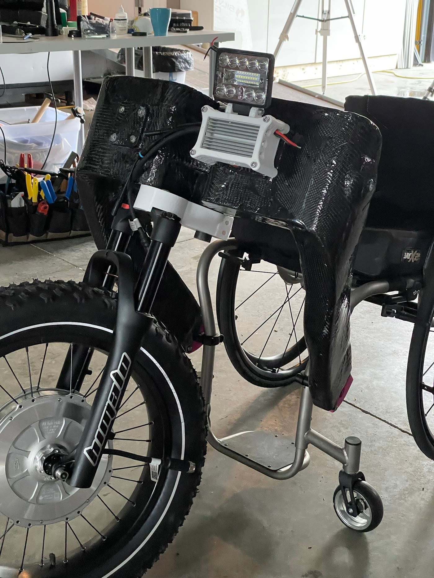 Carbon-fibre drive unit with suspension fork, headlight and printed mounts clamped to a manual wheelchair